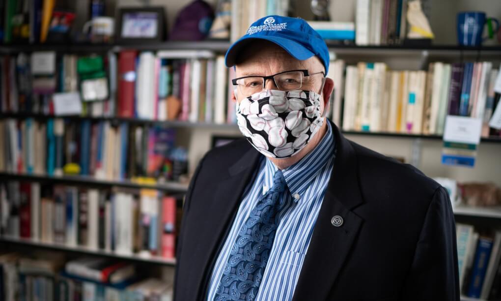 Fred Antczak standing in his office wearing a GVSU baseball cap and a mask with baseballs printed on it
